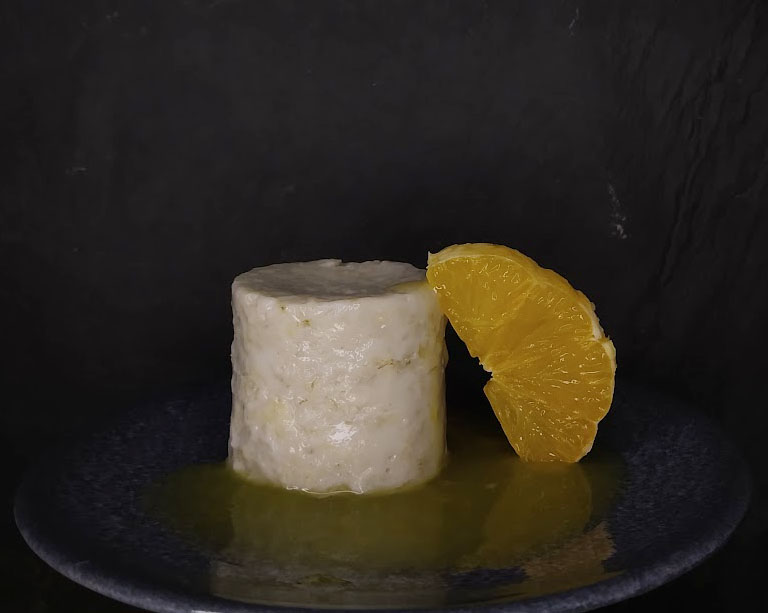 Auf dunklem Teller vor dunklem Hintergrund ein kleiner zylinderförmiger Porridge-"Pudding", daran gelehnt eine halbe Orangenscheibe.