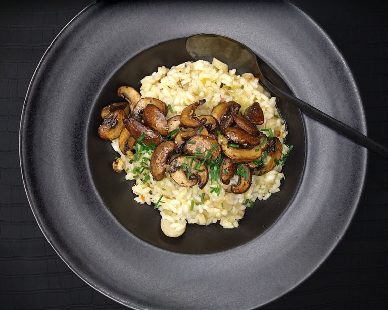 Dunkler Teller mit einer Portion hellem Risotto, quer darüber ein Streifen goldbraun gebratener Pilze.